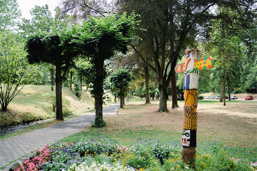 Im Vordergrund ist ein Blumenbeet zu sehen, in dem ein künstlerisch gestalteter Marterpfahl steht. Im Hintergrund ein Spazierweg, umsäumt von grünen Bäumen. Im Vordergrund ist ein Blumenbeet zu sehen, in dem ein künstlerisch gestalteter Marterpfahl steht. Im Hintergrund ein Spazierweg, umsäumt von grünen Bäumen.